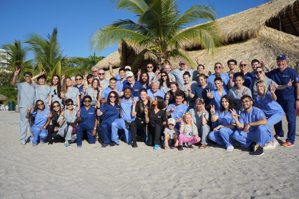 A large group in matching blue scrubs on a palm lined beach