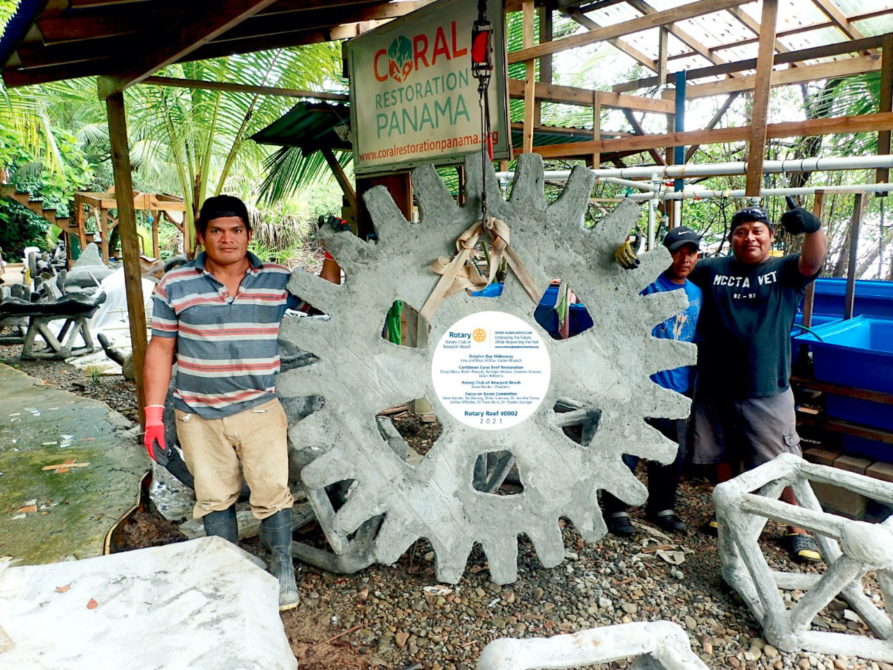 Two men beside a concrete reef module shaped like the Rotary wheel, in a workshop under a Coral Panama sign